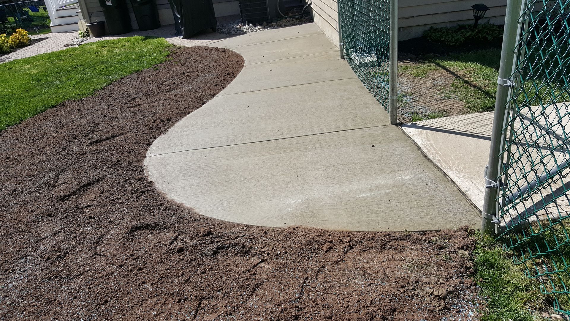 Curved concrete sidewalk beside mulched landscaping and a chain-link fence at a building entrance