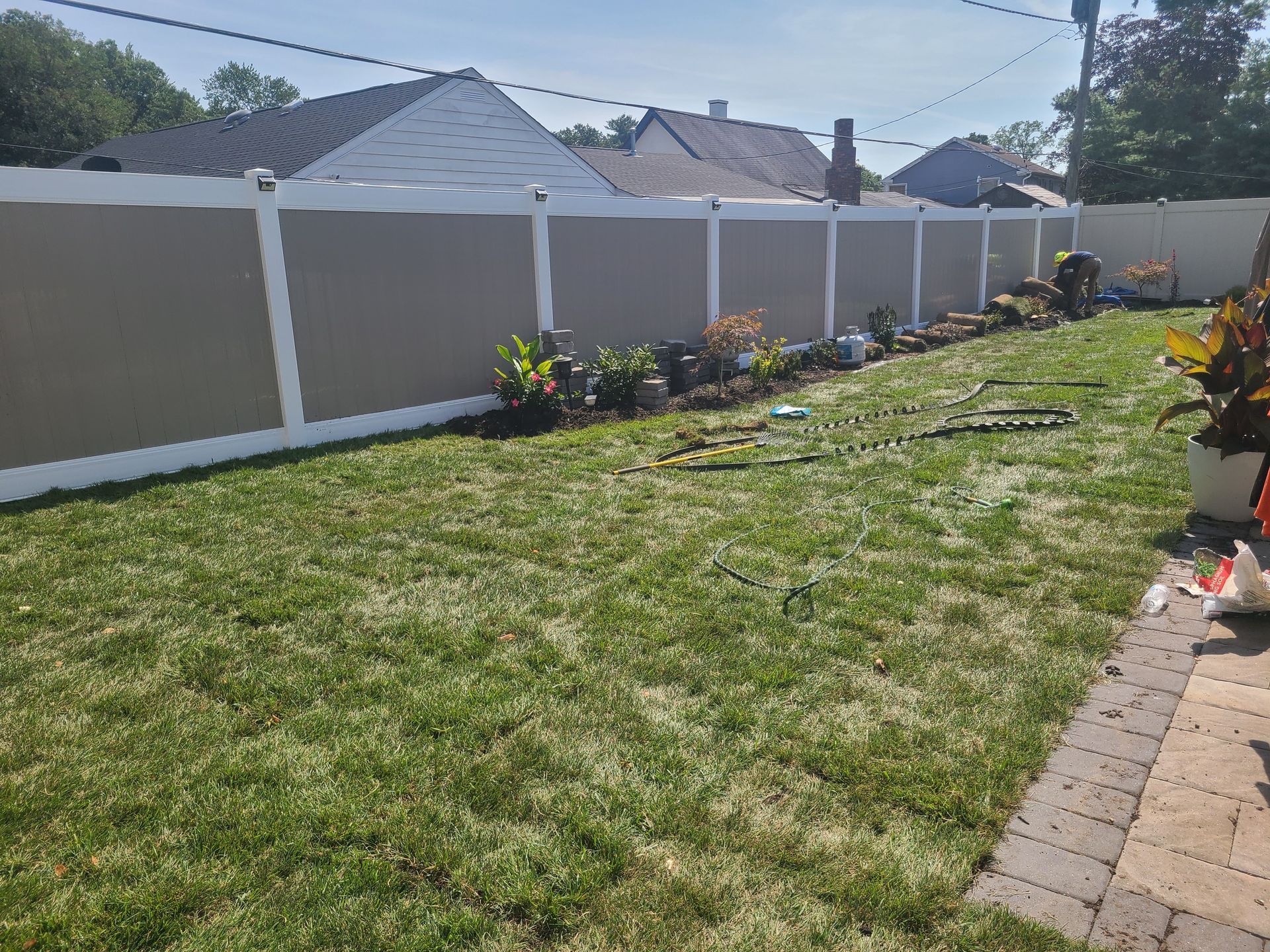 Backyard with patchy grass, white fence, and a brick patio along the right side under bright sunlight