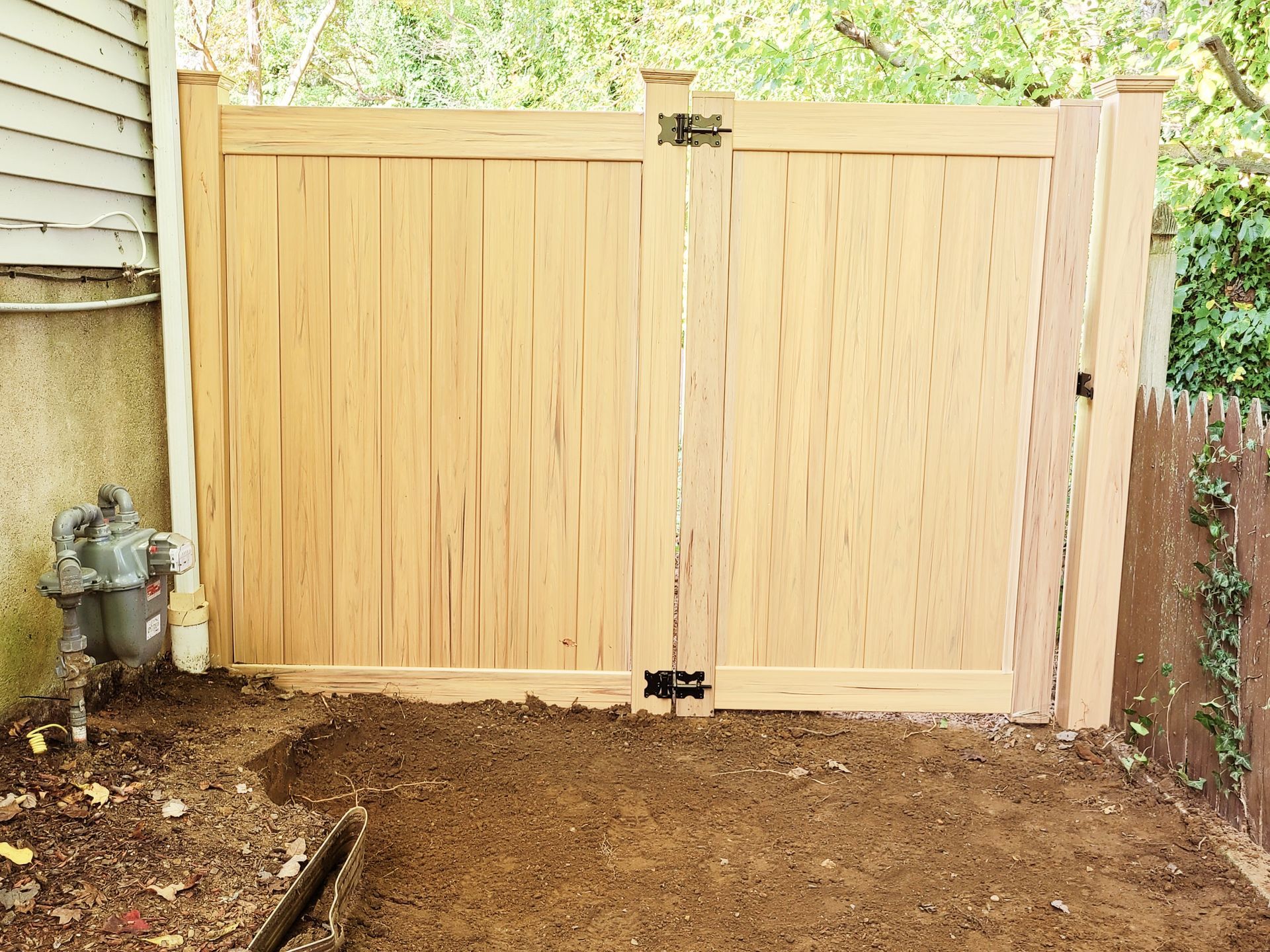 Light wood double gate closed across a dirt yard, with house siding and a brick wall on either side