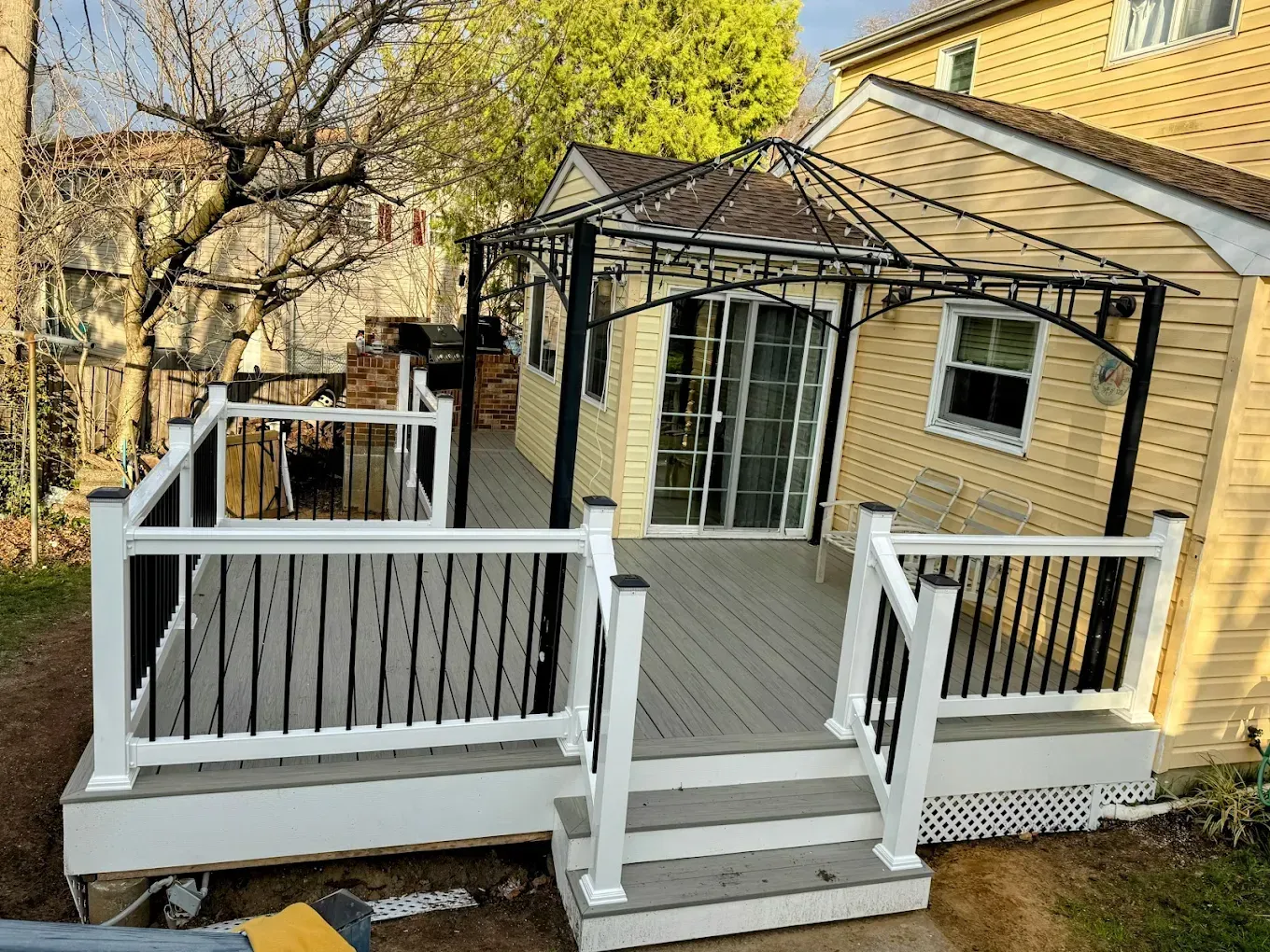 White wooden deck with steps and black metal pergola attached to a yellow house in a wooded backyard