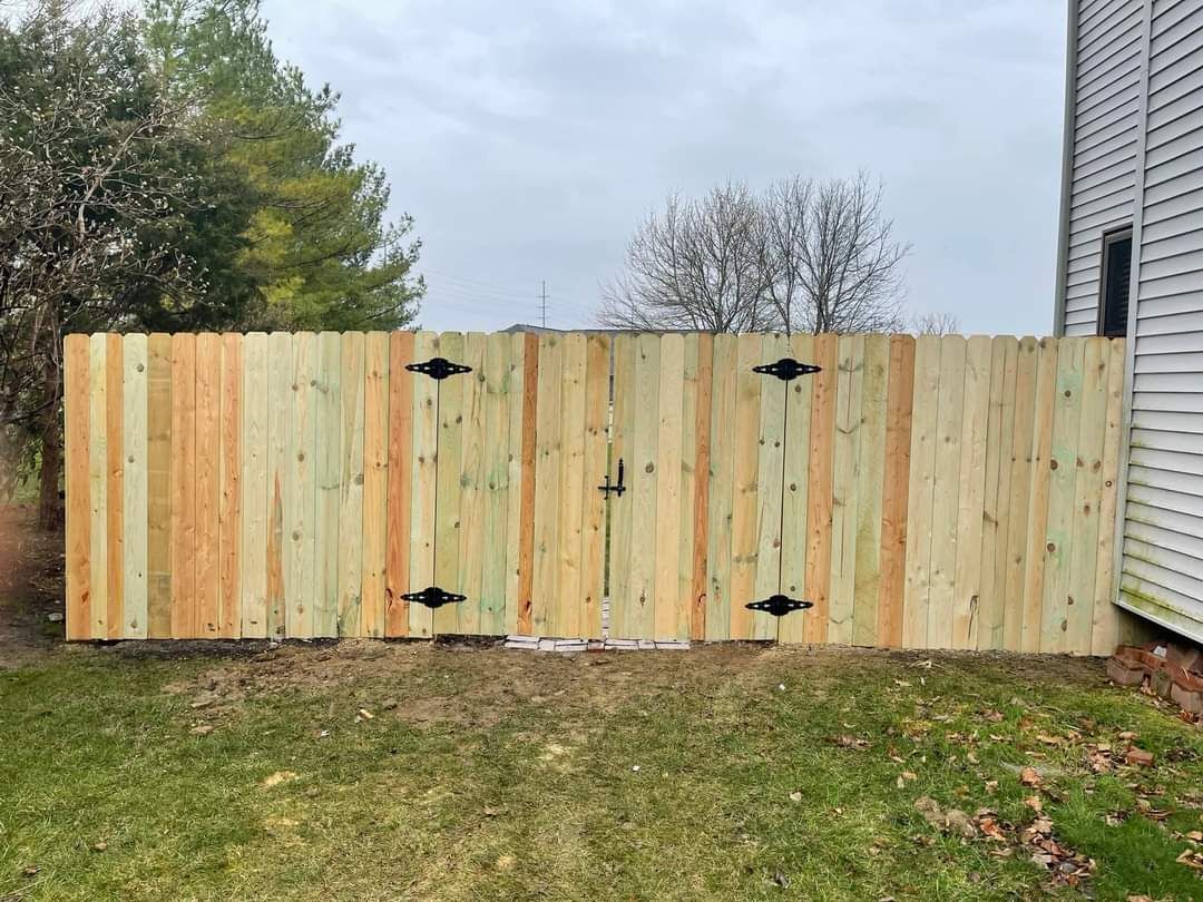 Wooden fence gates with black hinges beside a house, bordered by grass and trees