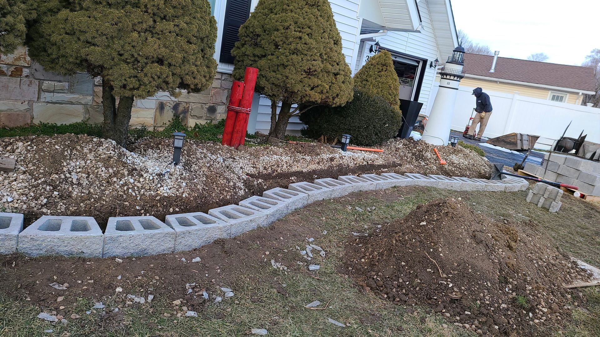 Curved stone border around a yard with gravel, mulch, trimmed trees, a red fire hydrant, and a worker near a house