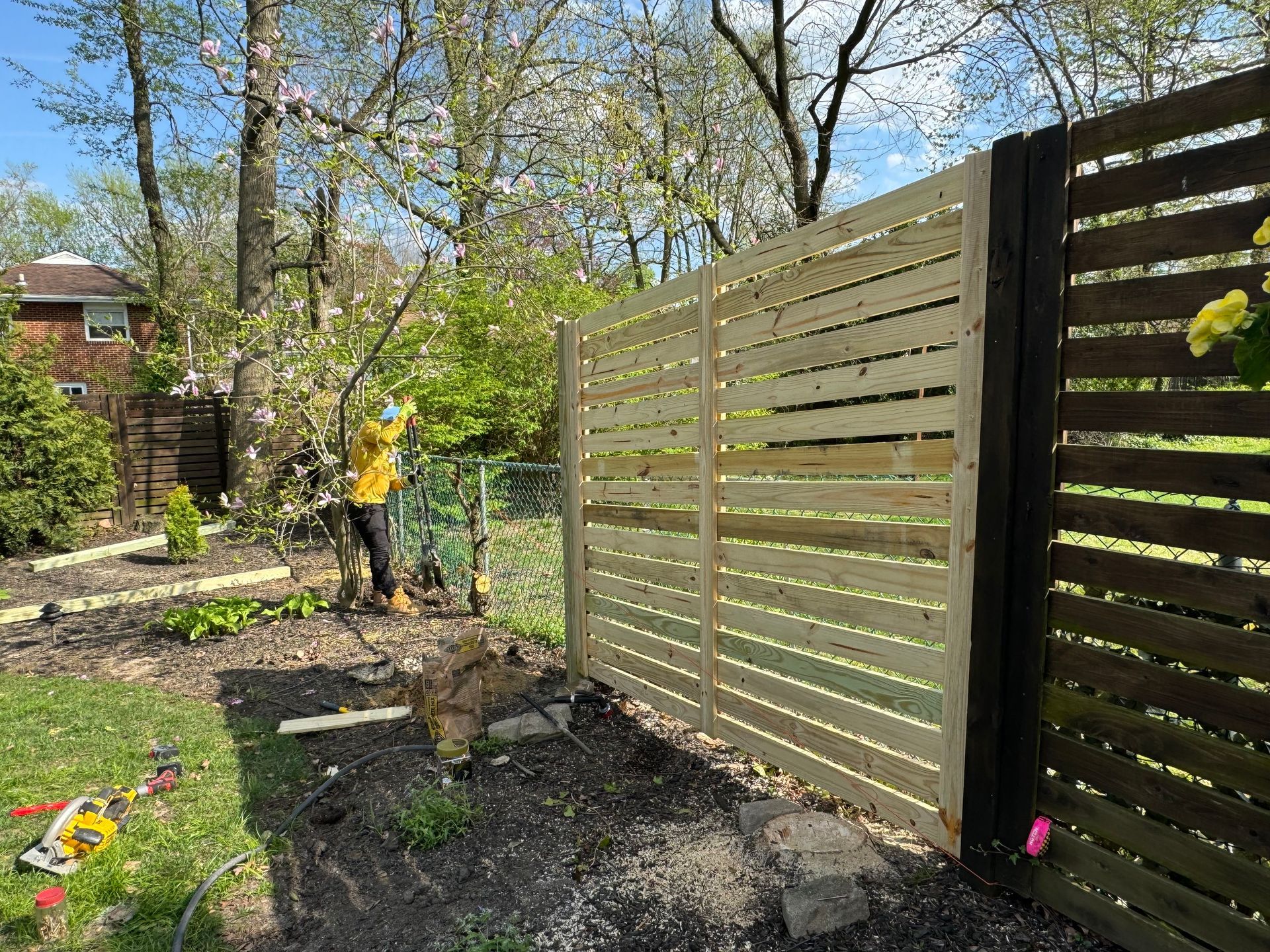 Person in yellow works in a garden beside a tan slat fence on a sunny day