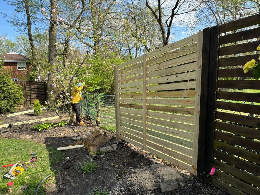 Person in yellow works in a garden beside a tan slat fence on a sunny day