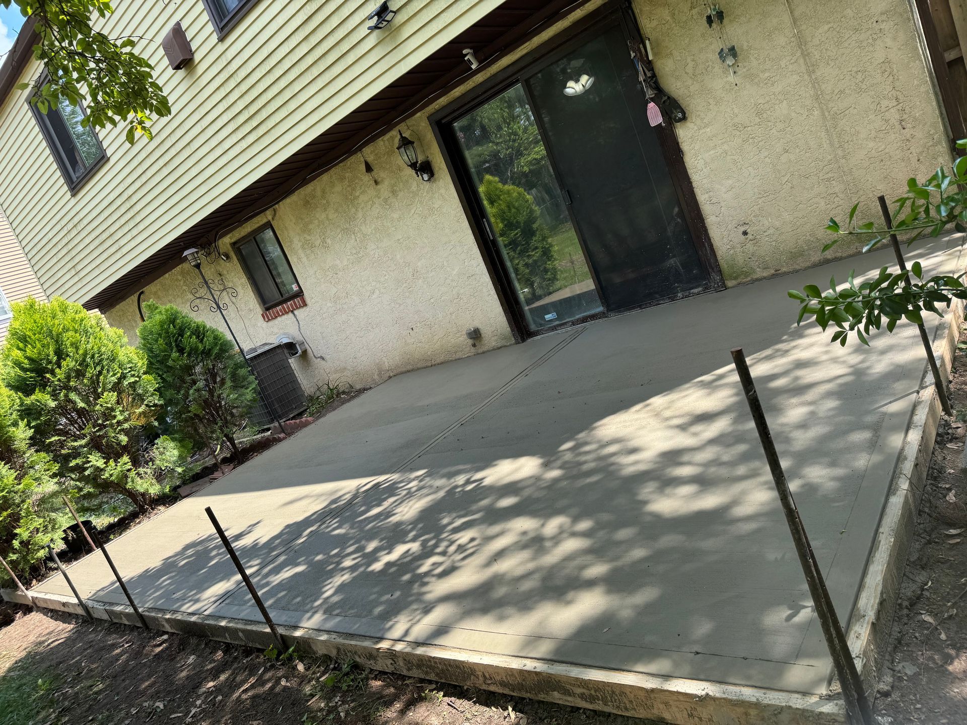 Concrete patio outside a house with a black door, beige walls, and shrubs along the left side.