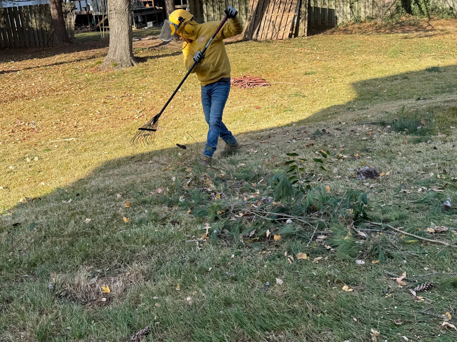 Person in a yellow hoodie rakes leaves in a grassy yard with trees and scattered autumn leaves.