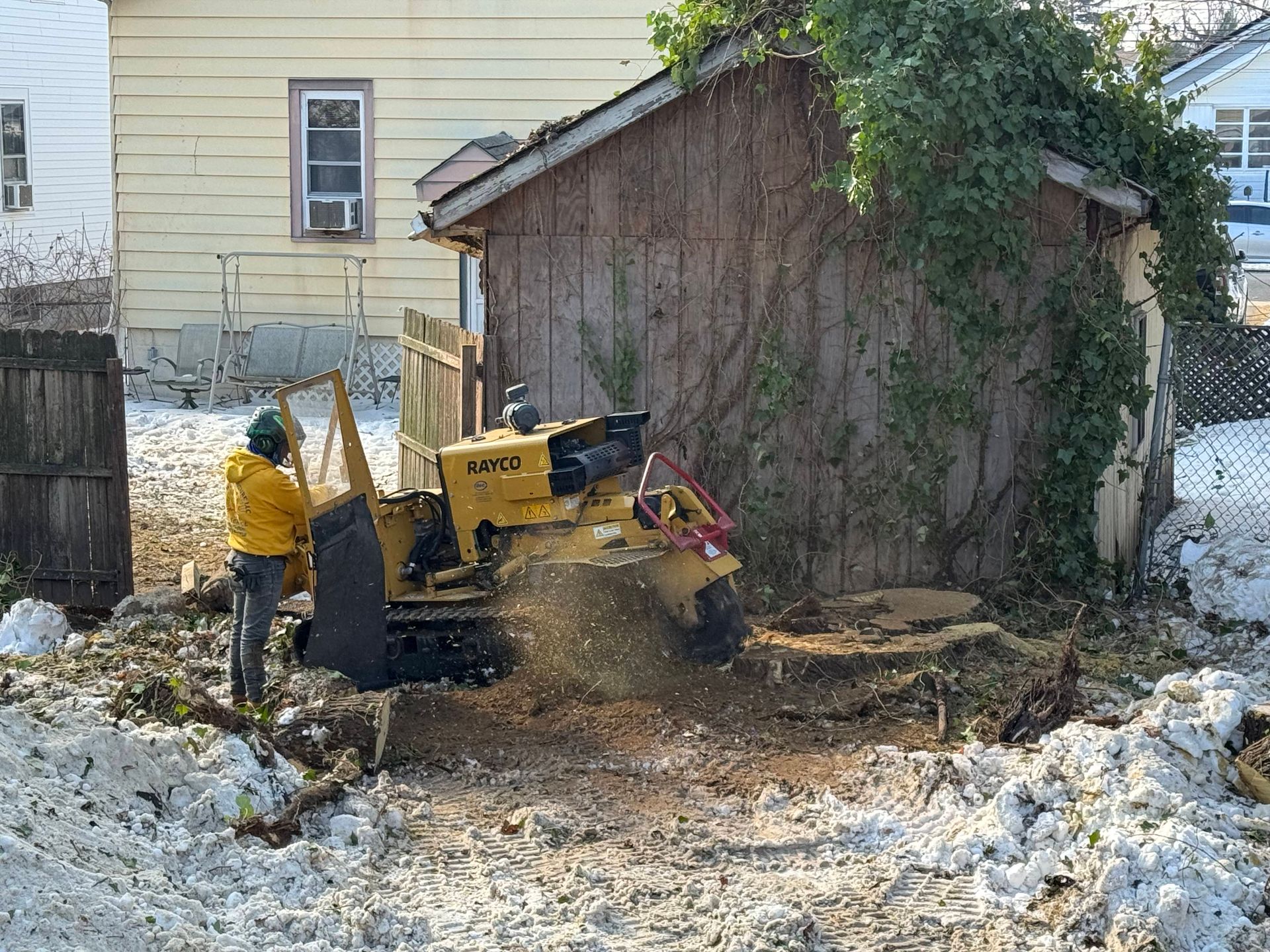 Person operating a yellow tractor in a snowy backyard beside a shed