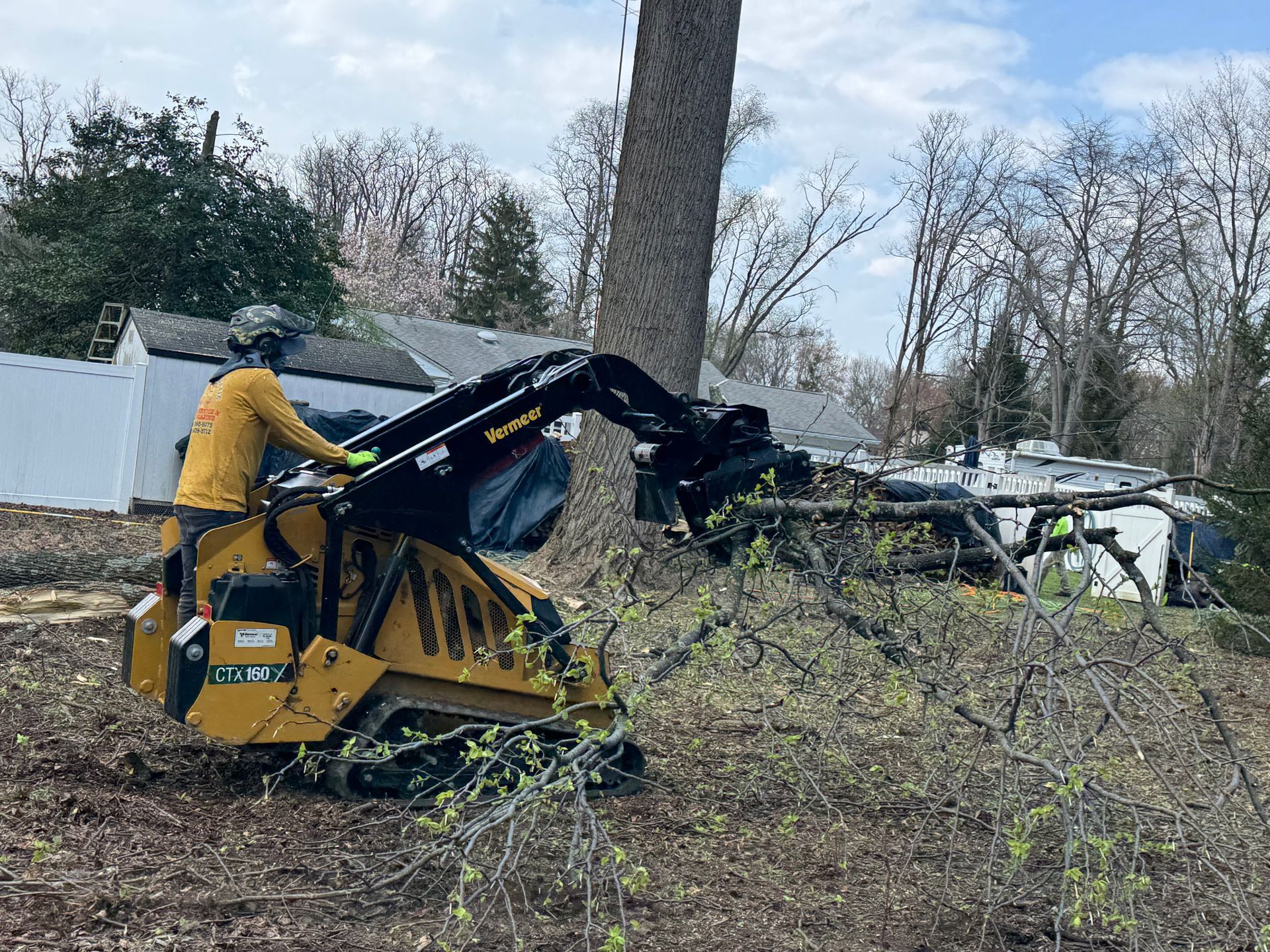 Person in yellow jacket operating a yellow skid steer clearing brush in a yard with trees and a white fence