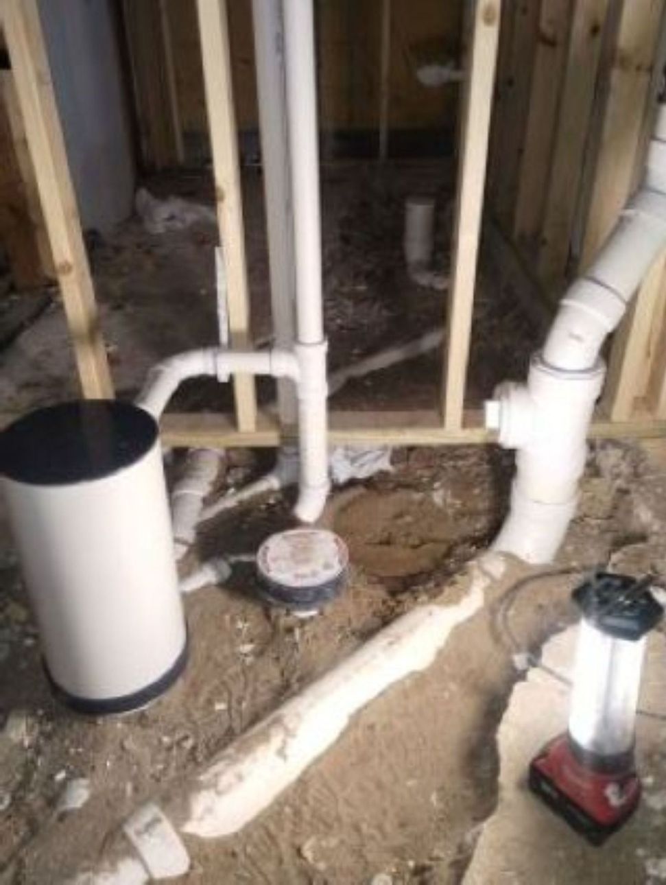 Plumbing under construction in a crawl space, with white PVC pipes, a drain, and a flashlight.