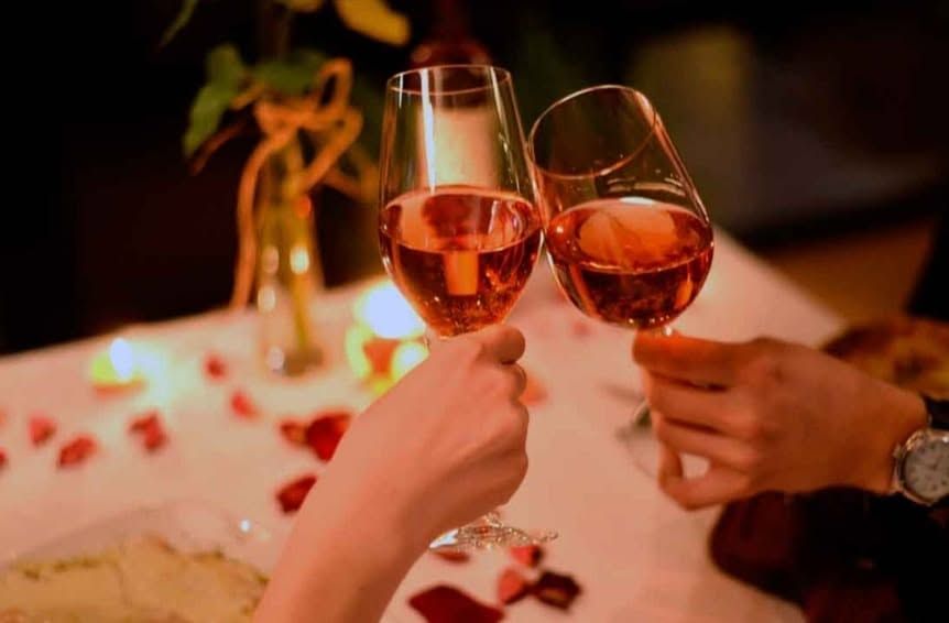Dos personas brindan con copas de vino rosado en una mesa iluminada con velas, adornada con pétalos de rosa y flores.