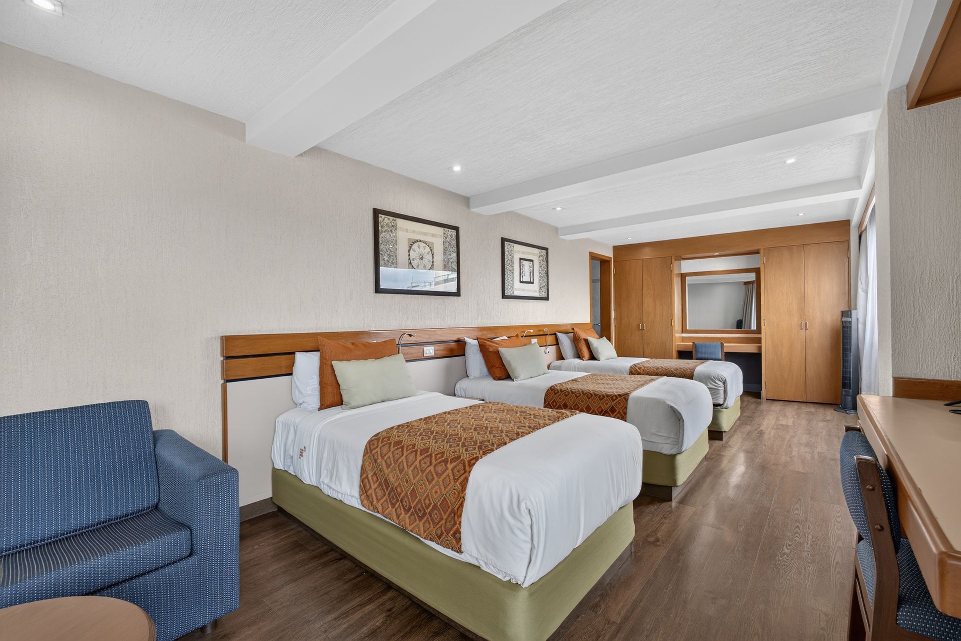Habitación de hotel con tres camas bien hechas, paredes beige y cuadros paisajísticos enmarcados.