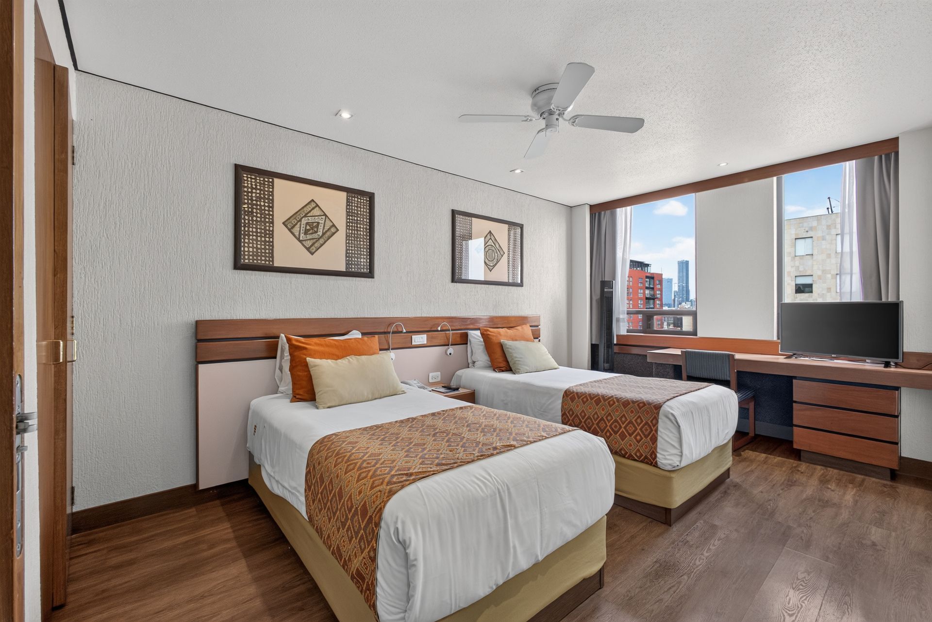 Habitación de hotel con dos camas individuales, ropa de cama beige, cabeceros de madera y una gran ventana con cortinas.