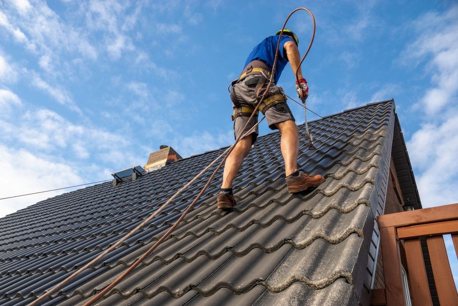 Roof worker wearing a harness climbs a steep dark-tiled roof against a blue sky