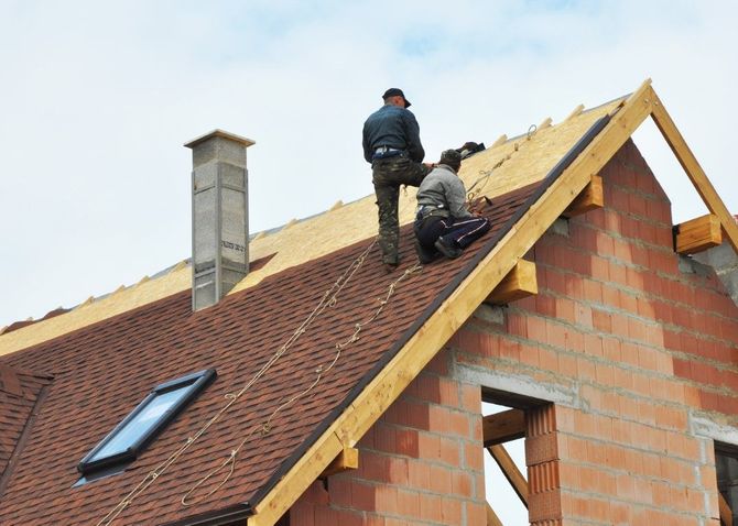 Two workers installing shingles on a roof of a brick house