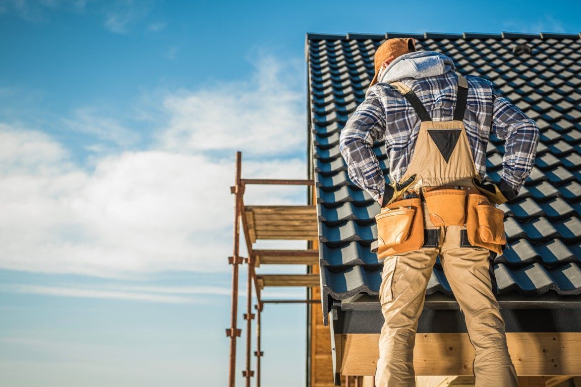 Worker on a rooftop ladder beside blue roof tiles under a clear sky