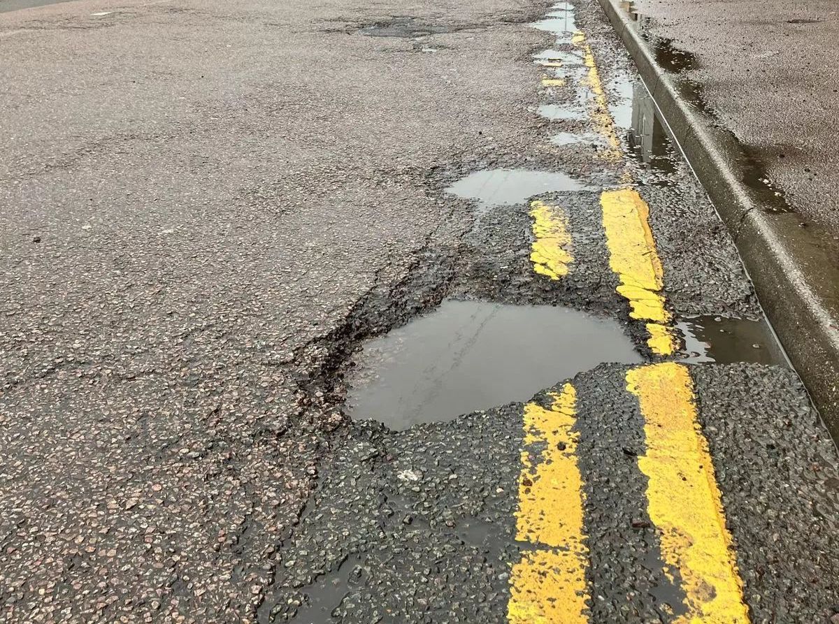 Potholes filled with water on a road with faded yellow lines, next to a curb.