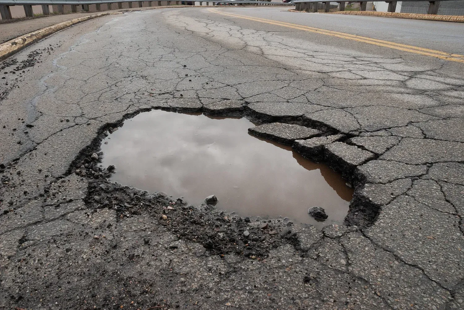 Pothole filled with water on cracked asphalt road. Sky reflection visible.