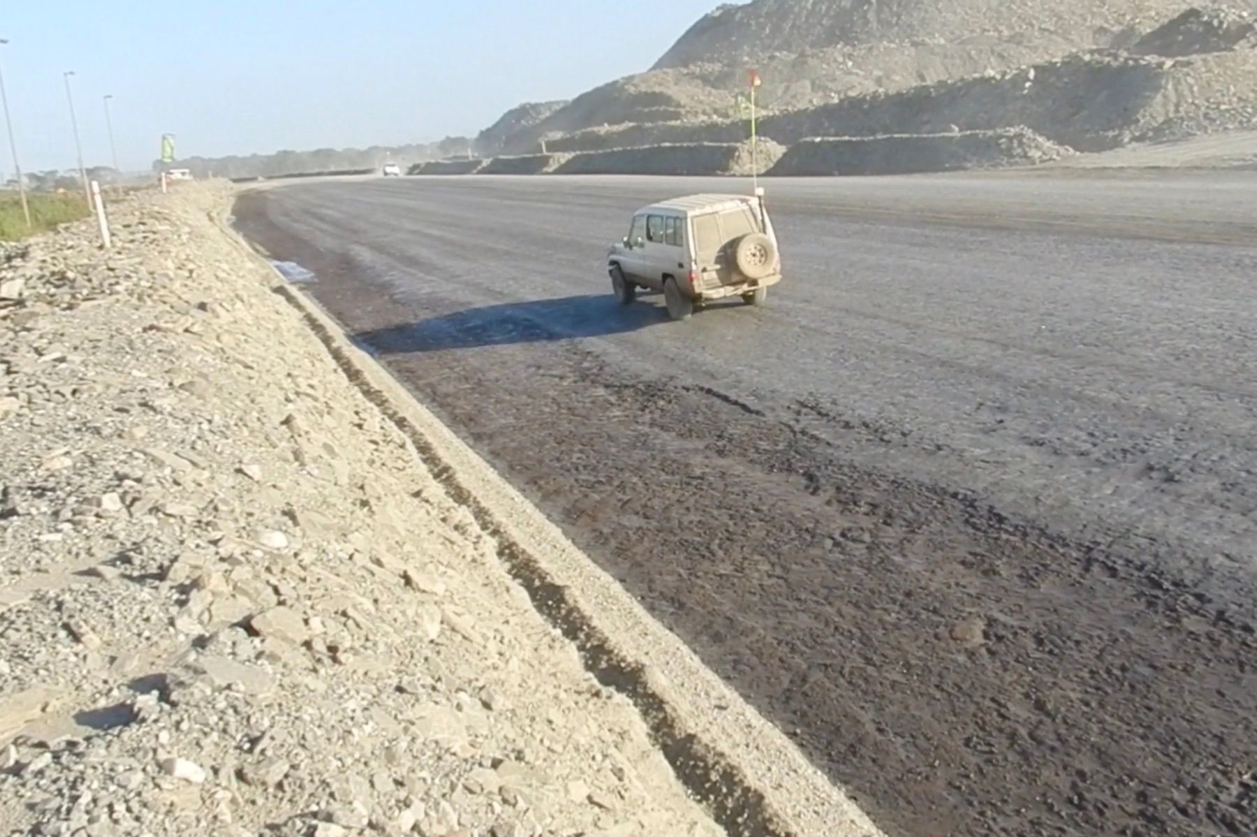 An SUV drives on a freshly paved road, with a rocky hillside and dust in the background.