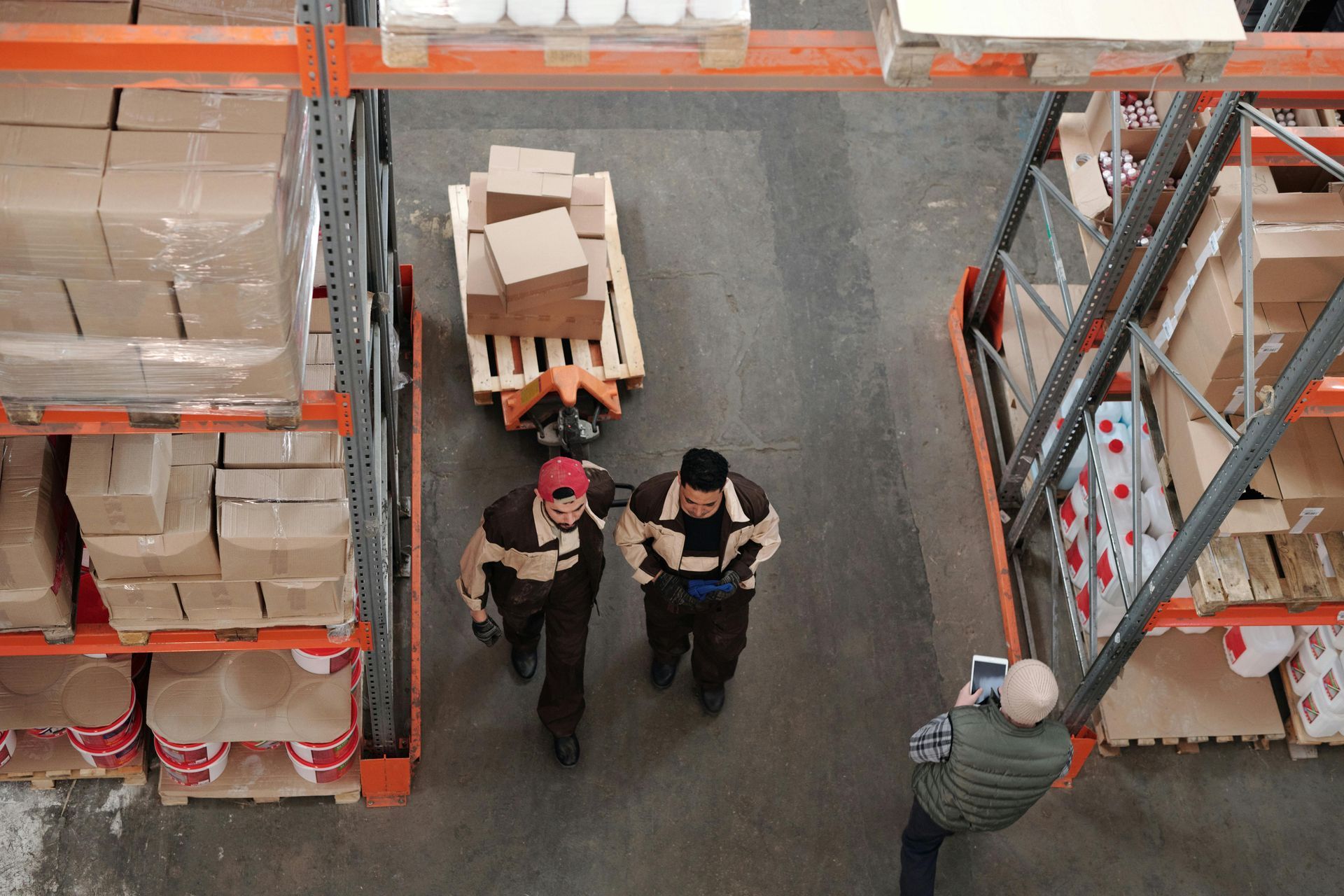 Workers in warehouse, pushing a cart with boxes. Another person is taking a photo. Shelving units on sides.