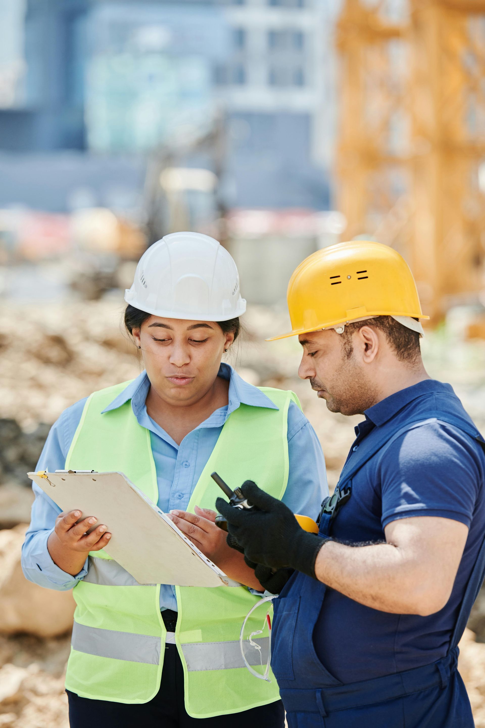 Construction workers reviewing documents on site. One wears a white hard hat and vest, the other a yellow hard hat.