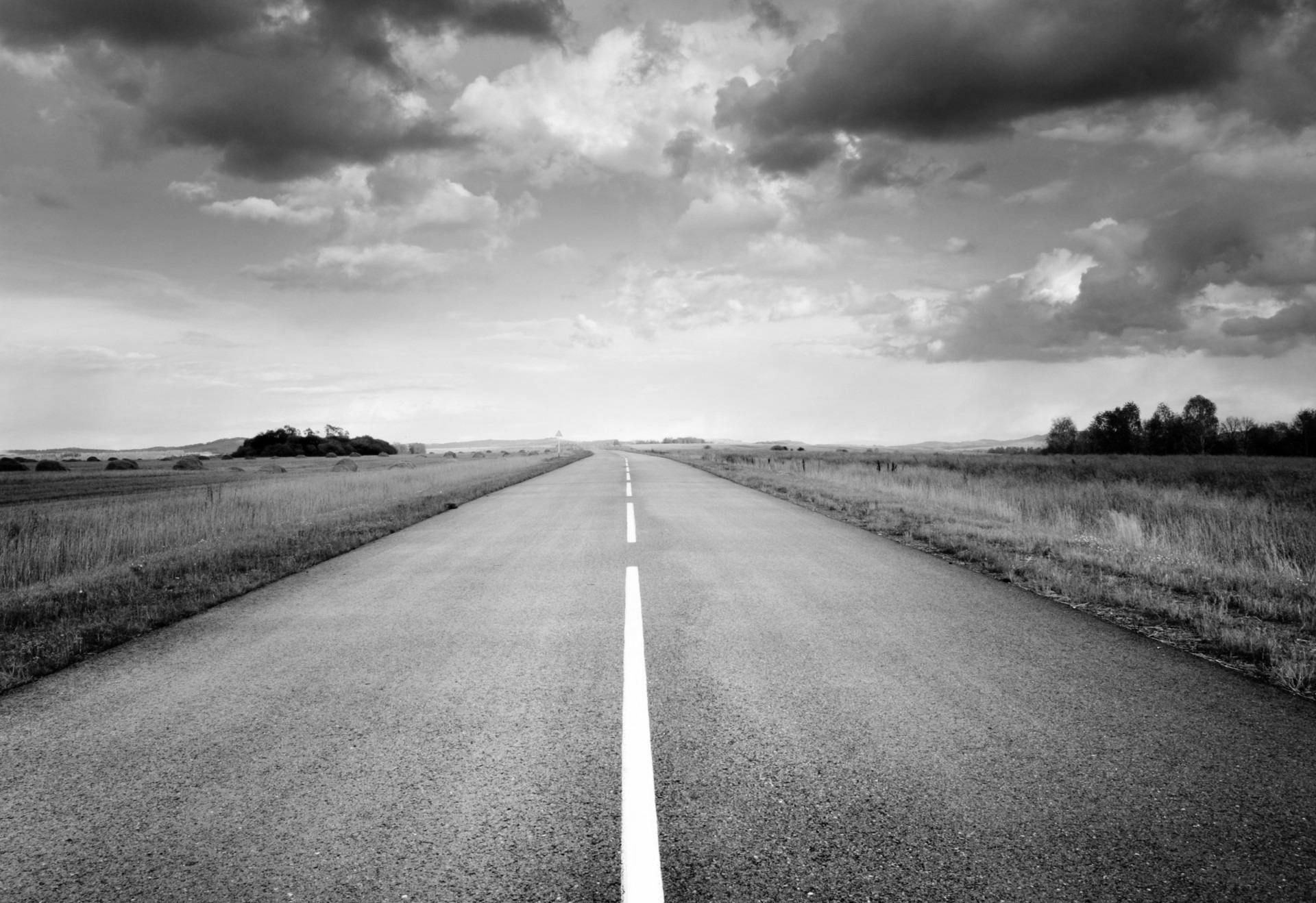 Black and white photo: a long, straight road stretches toward the horizon, under a cloudy sky.