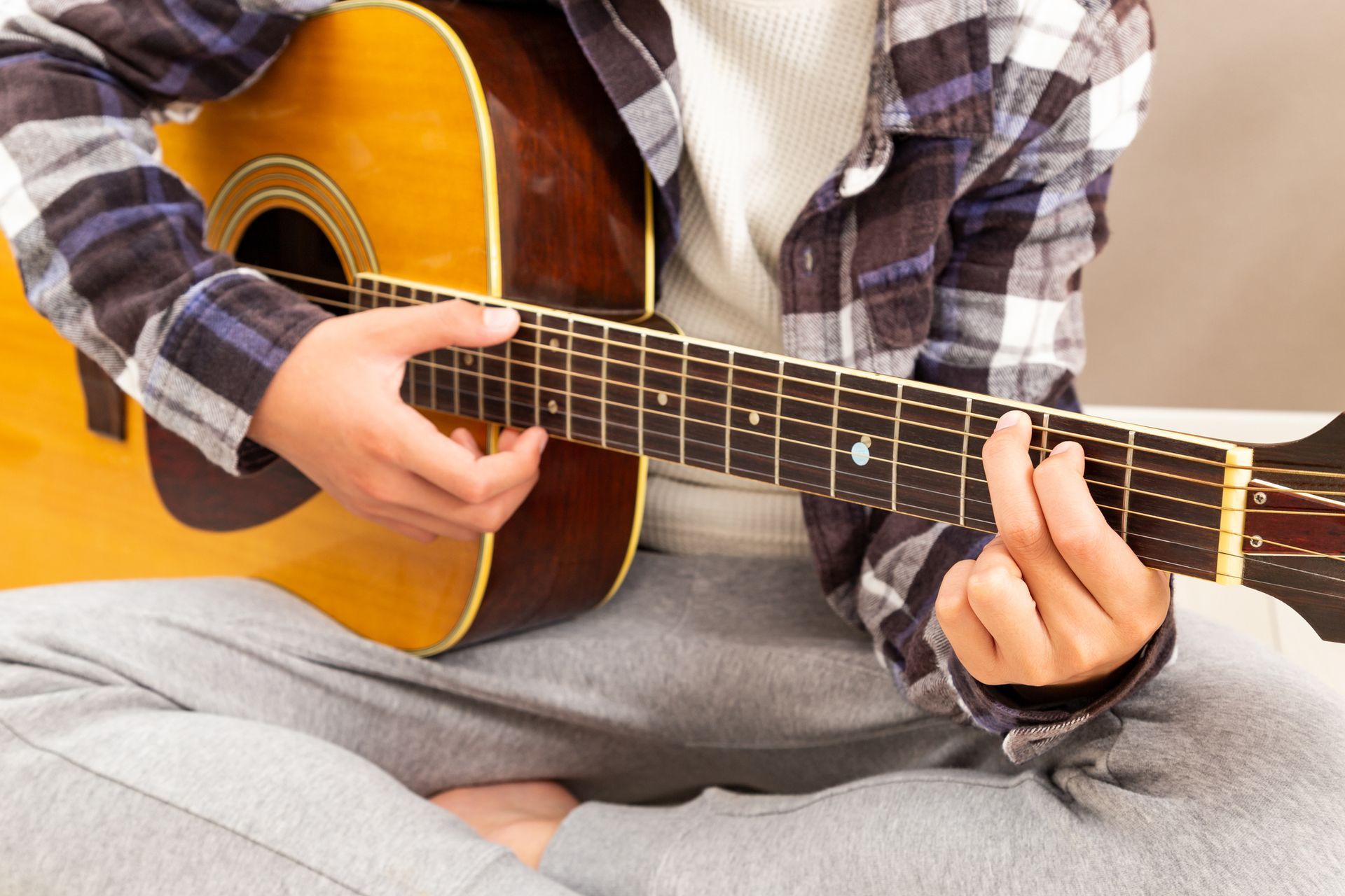 Person playing acoustic guitar, sitting cross-legged. Wearing plaid shirt, grey sweats.