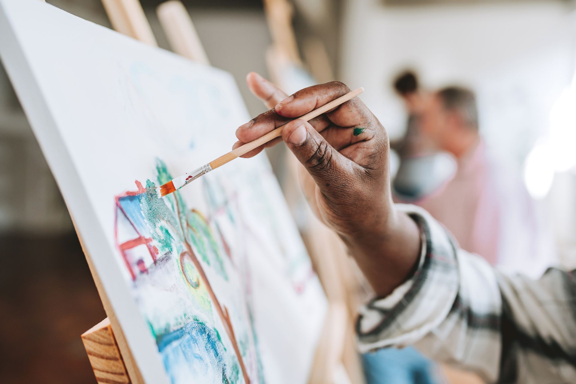 Person painting with brush on canvas, depicting house and trees, with others in background.