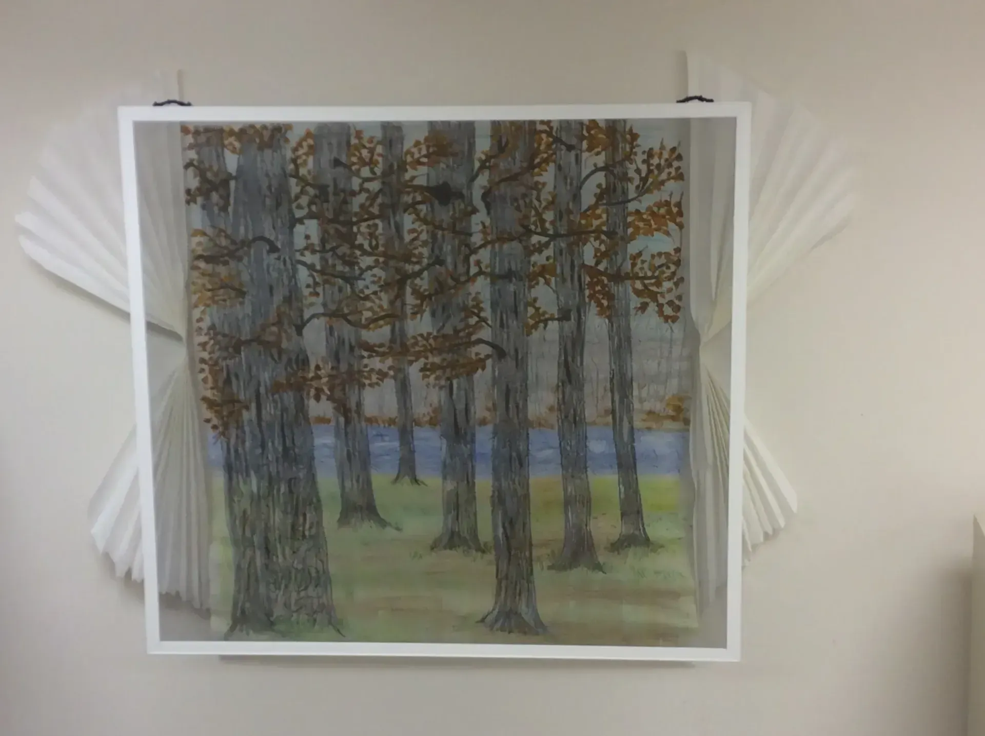 Watercolor painting of trees by a lake, framed with white paper art on a white wall.