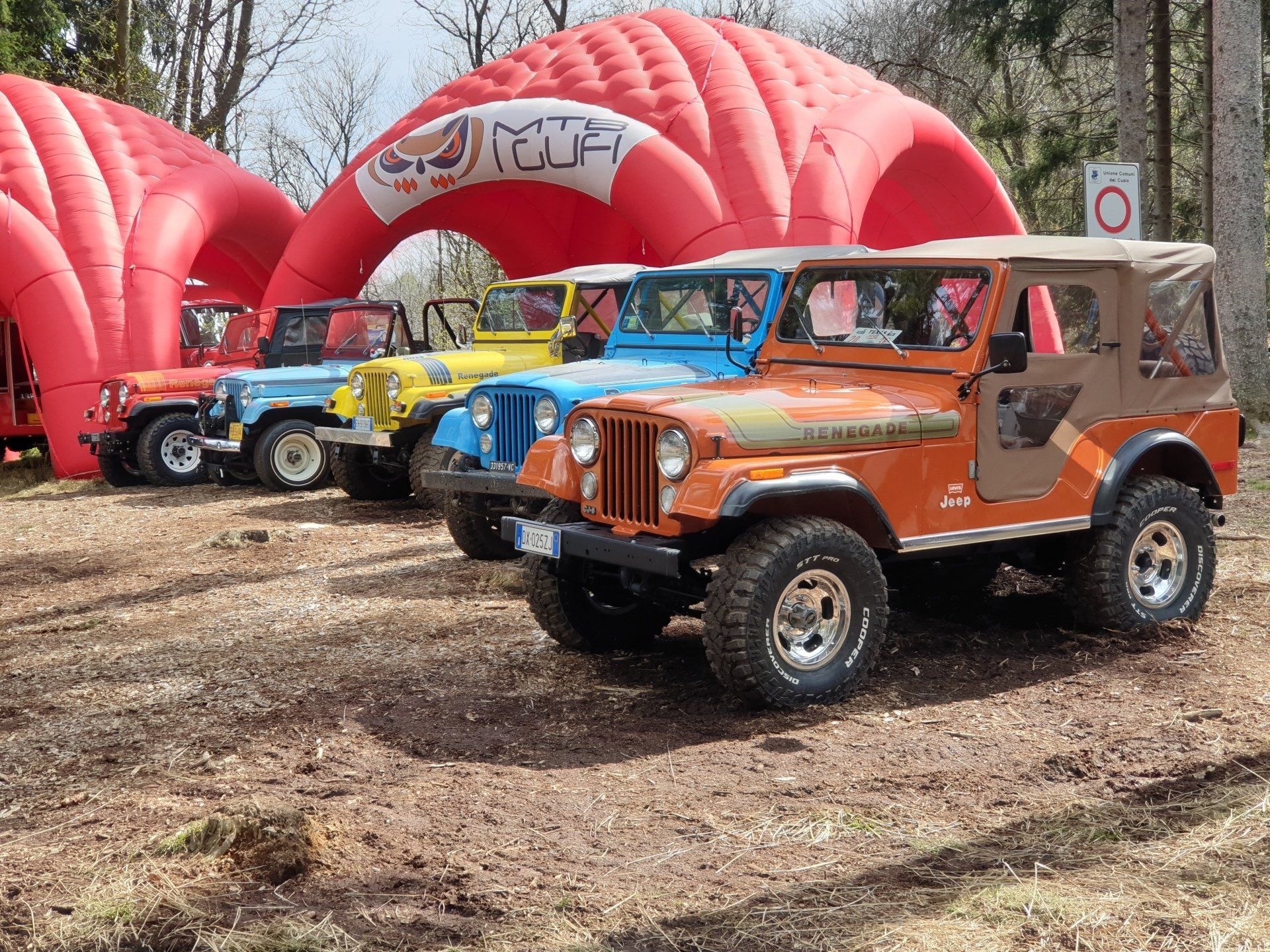 Cinque Jeep Wrangler d'epoca parcheggiate in un bosco, con un arco gonfiabile rosso sullo sfondo.