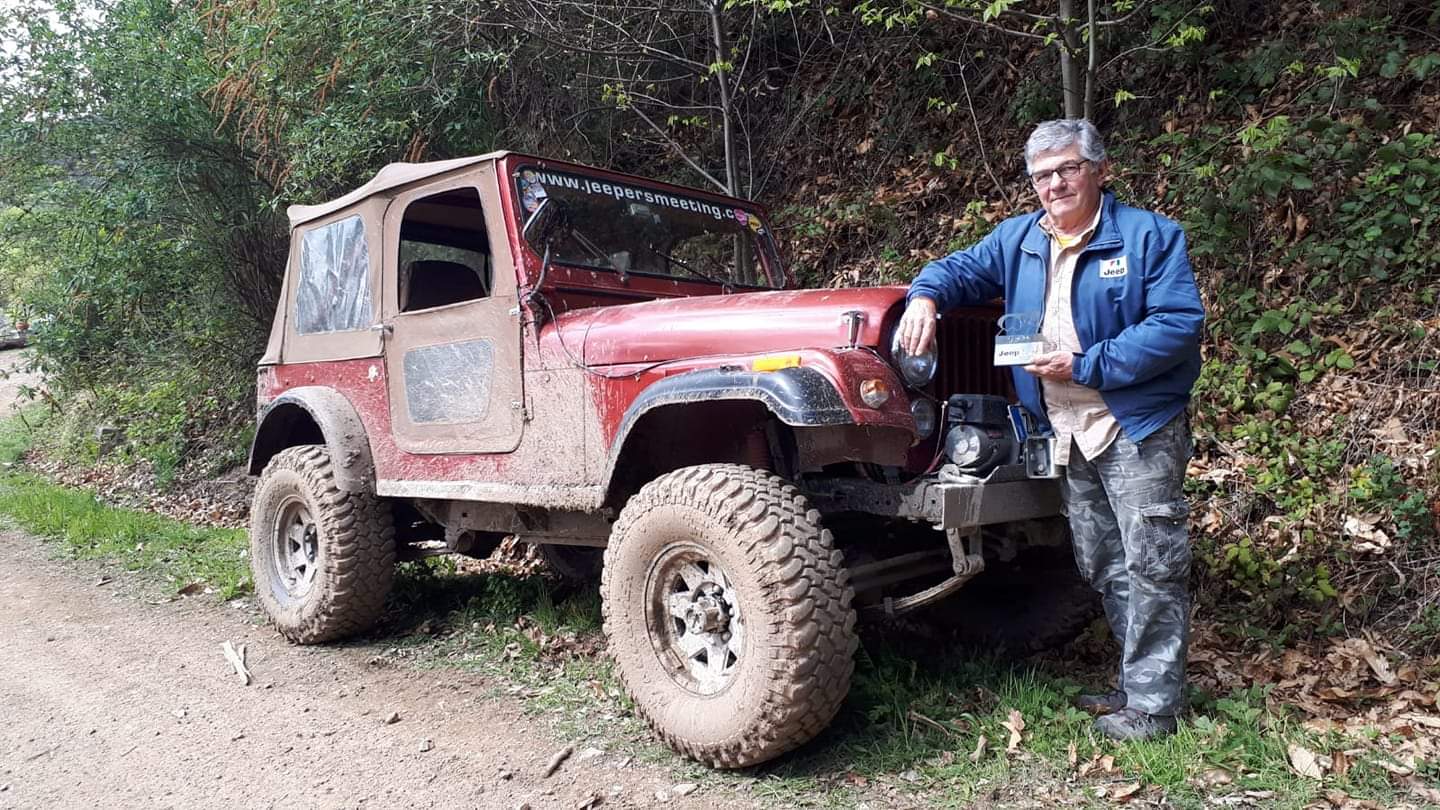 Un uomo in piedi accanto a una Jeep rossa infangata su una strada sterrata tra gli alberi.