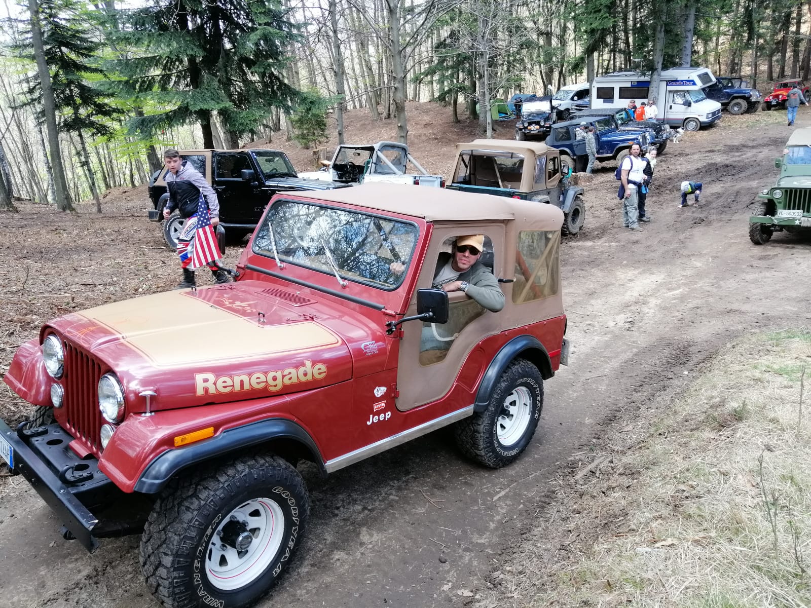 Una Jeep Renegade rossa guida una fila di veicoli fuoristrada su un sentiero sterrato in una zona boscosa; il conducente fa segno con la mano.