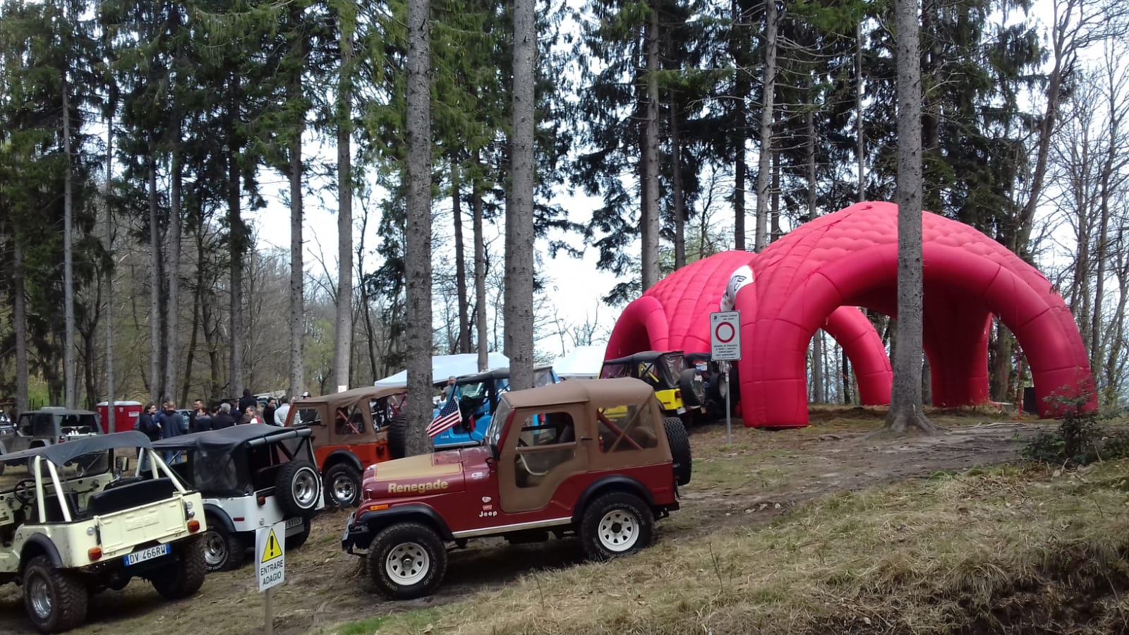 Jeep parcheggiate in una zona boscosa, vicino a una struttura gonfiabile rossa. La gente si raduna lì vicino.
