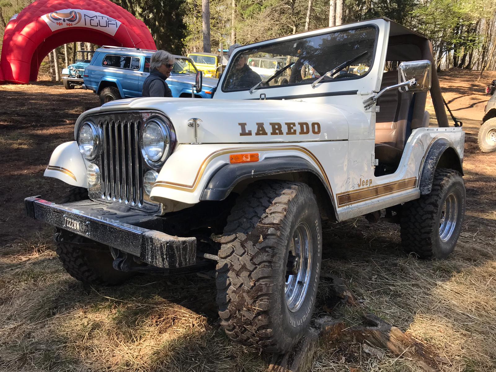 Jeep Laredo bianca, con dettagli marroni e dorati, parcheggiata all'aperto insieme ad altri veicoli; sullo sfondo è visibile un arco rosso.