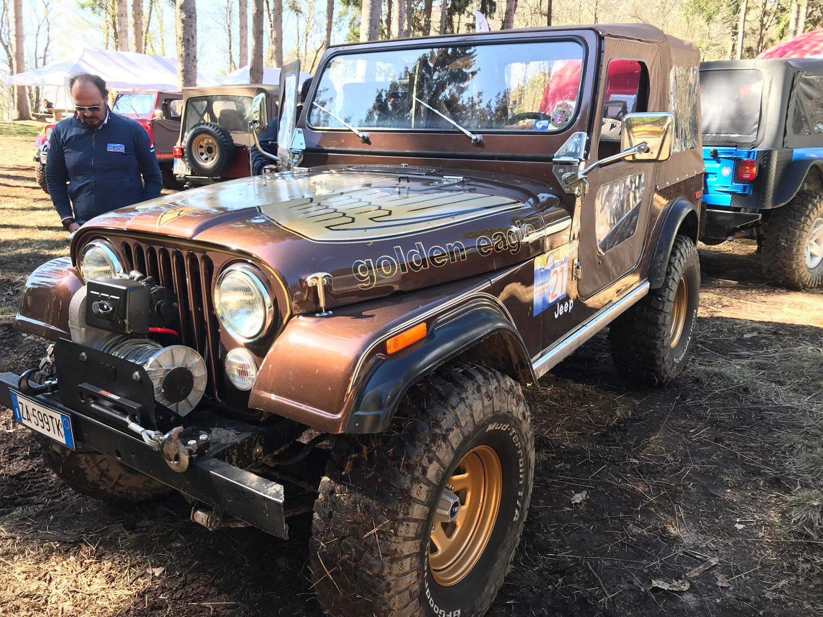 Una Jeep marrone con cerchi dorati, in un ambiente fangoso, con un uomo che la osserva.