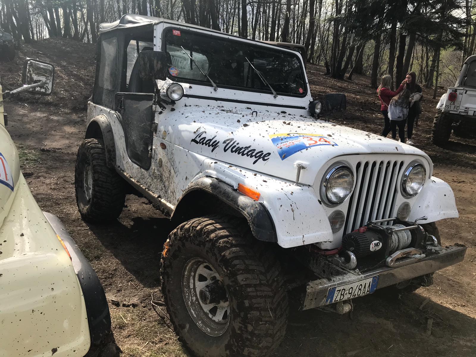 Jeep bianca d'epoca con fango, parcheggiata su una strada sterrata in una zona boscosa; scritta 