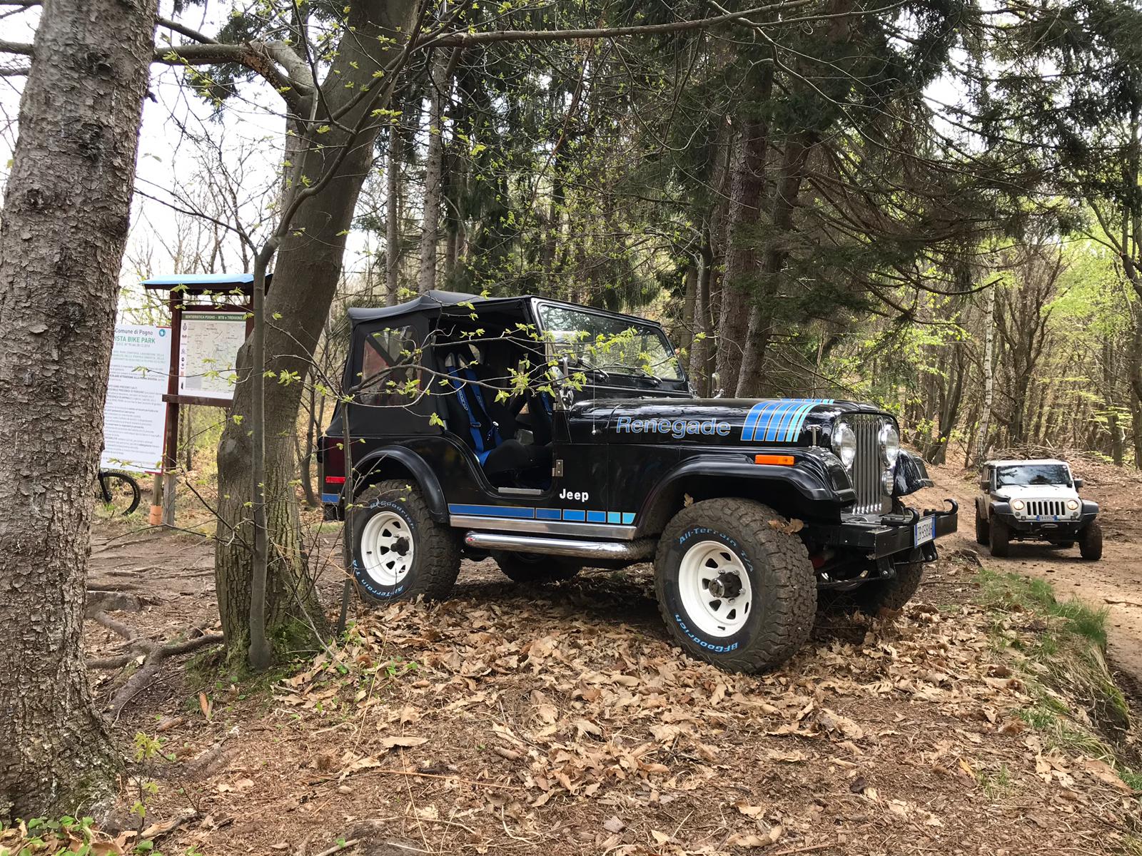 Jeep nera e blu parcheggiata su un sentiero alberato. Veicolo robusto con ruote bianche vicino a un cartello. Un'altra Jeep sullo sfondo.