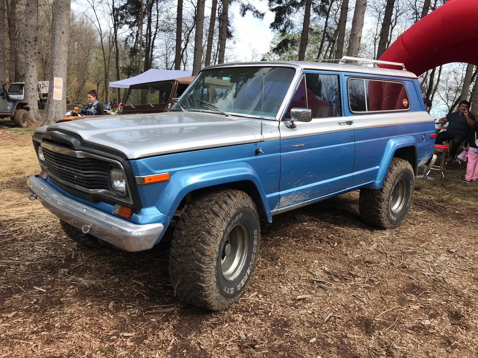 Veicolo fuoristrada Jeep blu e argento con pneumatici di grandi dimensioni, parcheggiato su terra con persone e alberi sullo sfondo.