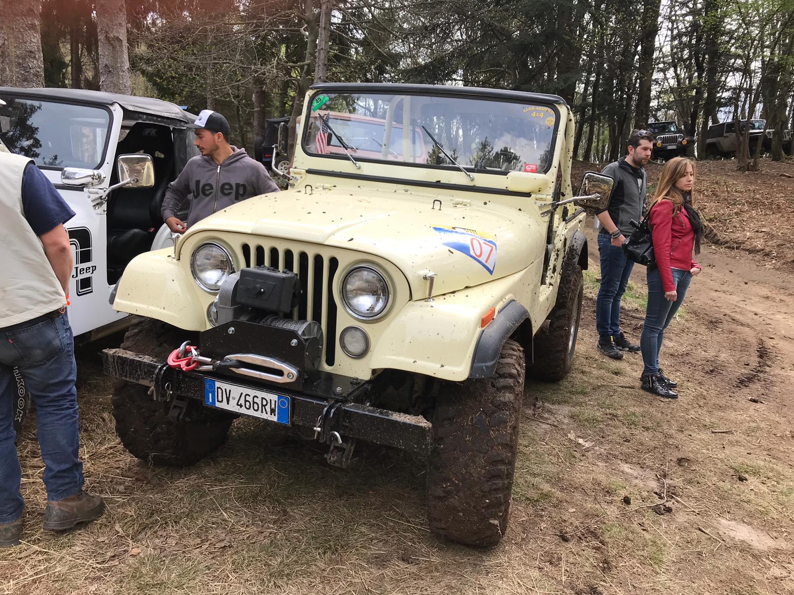 Jeep beige con pneumatici infangati, parcheggiata sullo sterrato. La gente è in piedi intorno ad essa in una zona boscosa.