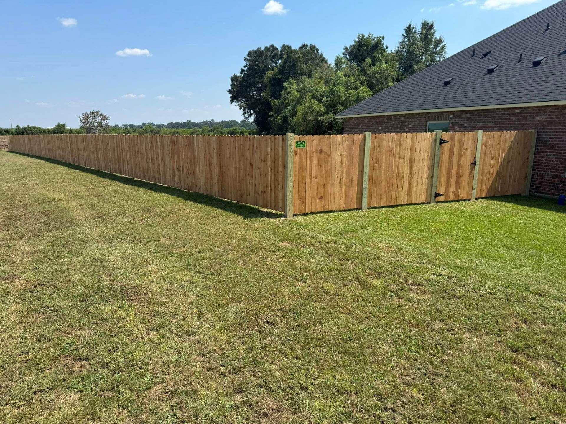 Wooden fence along a grassy yard, beside a brick house on a sunny day.