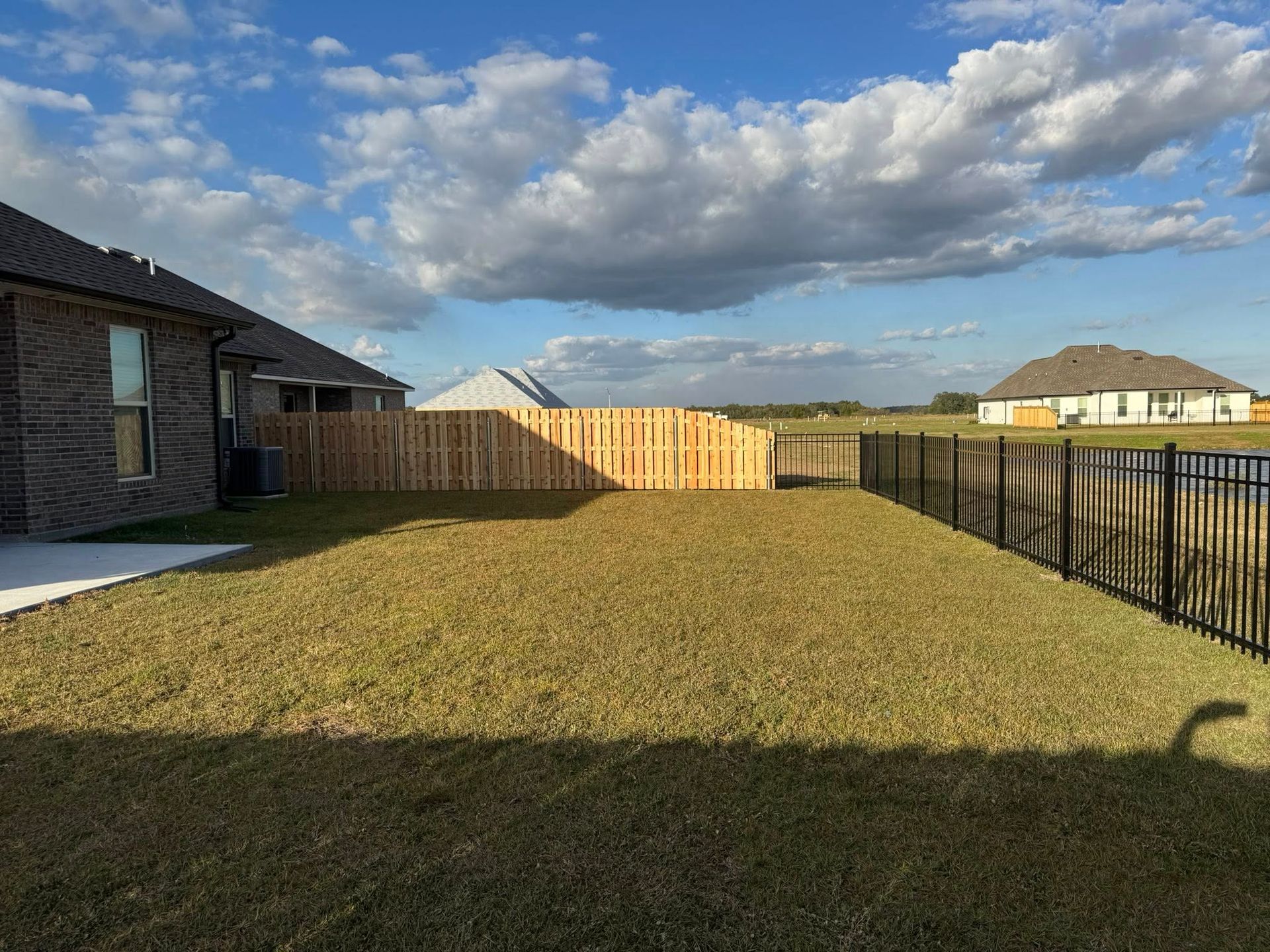 Grassy backyard with wooden fence, black metal fence, and two houses on a sunny day with clouds.