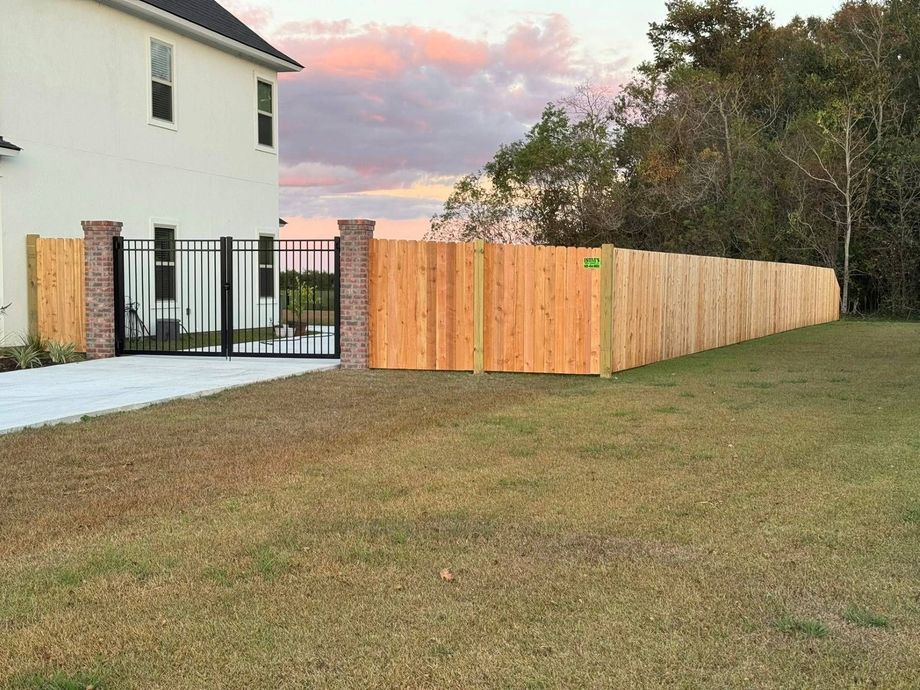 Wooden fence surrounding a grassy yard next to a white house with a black gate.