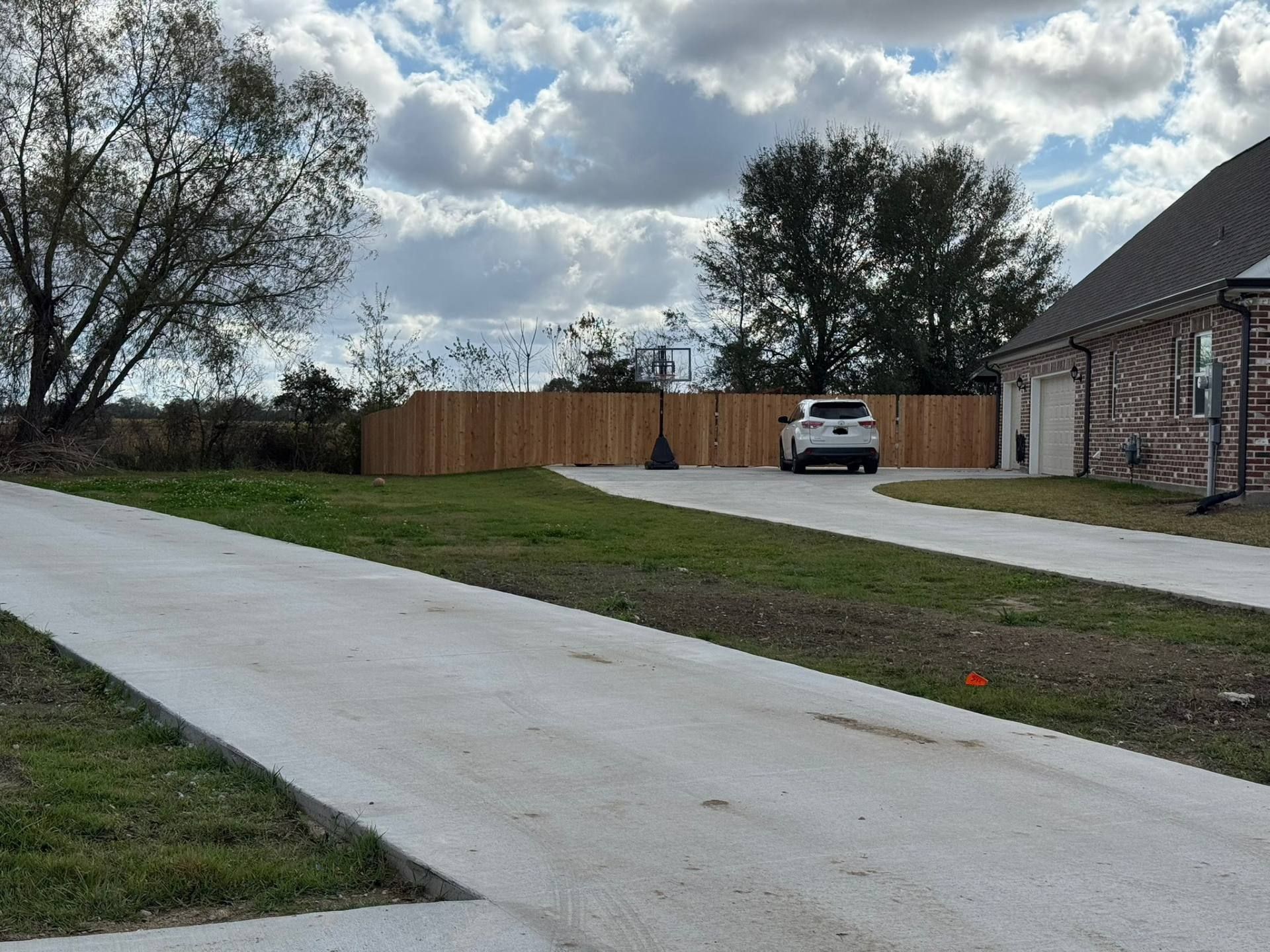 Driveway leads to a house with a car parked in front of a wooden fence; cloudy sky.