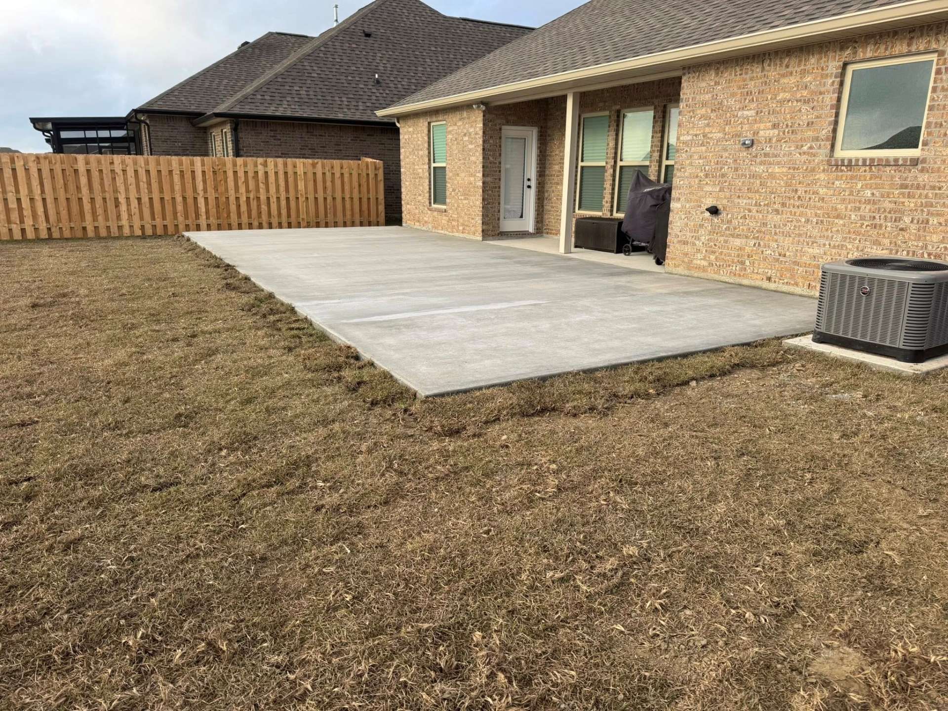 Concrete patio in backyard with brick house and brown grass.