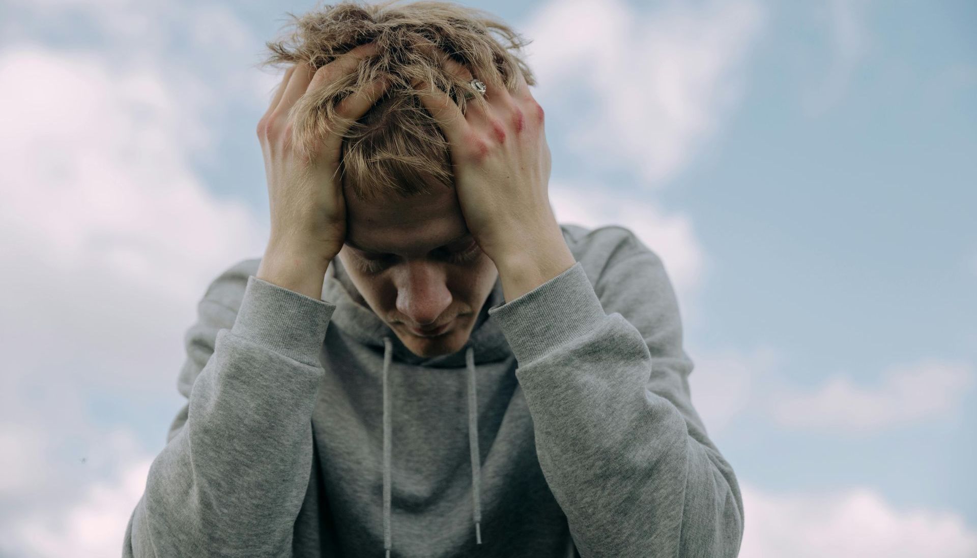 A man is holding his head in his hands in front of a cloudy sky.