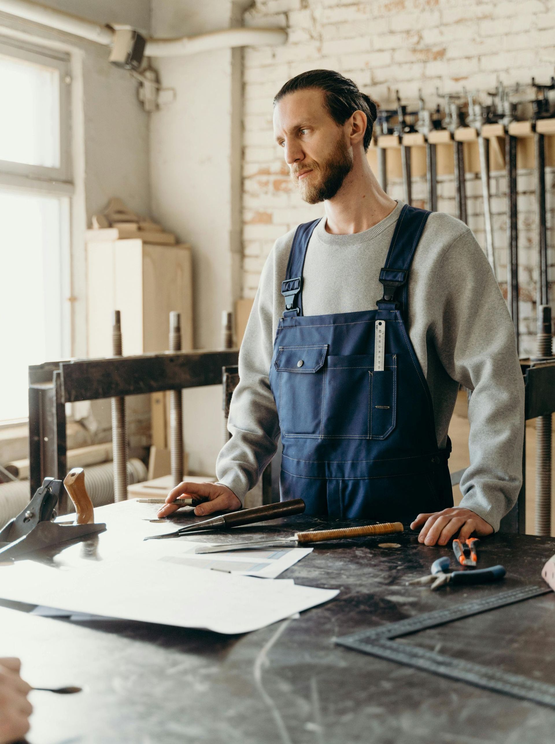 A man in overalls is standing at a table in a workshop.