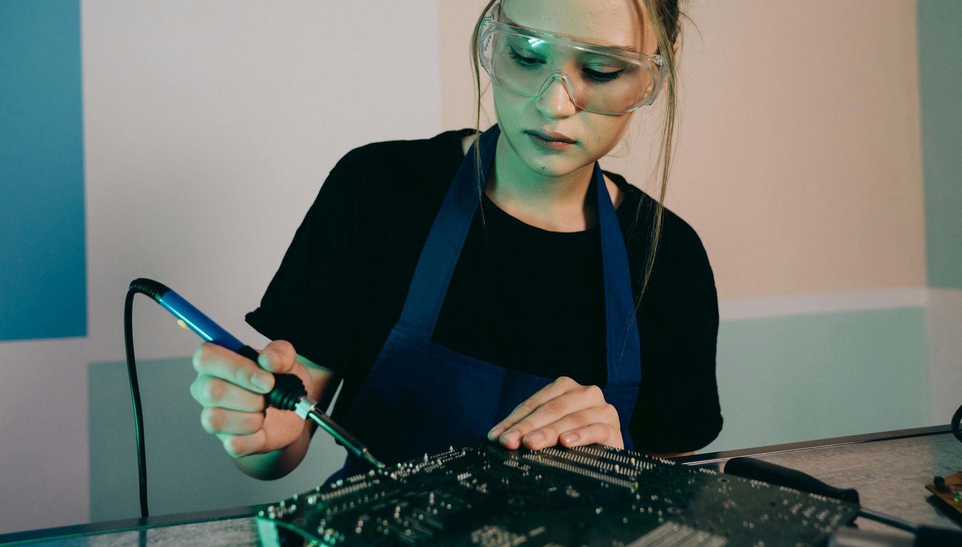 A woman is wearing safety glasses and an apron while working on a motherboard.
