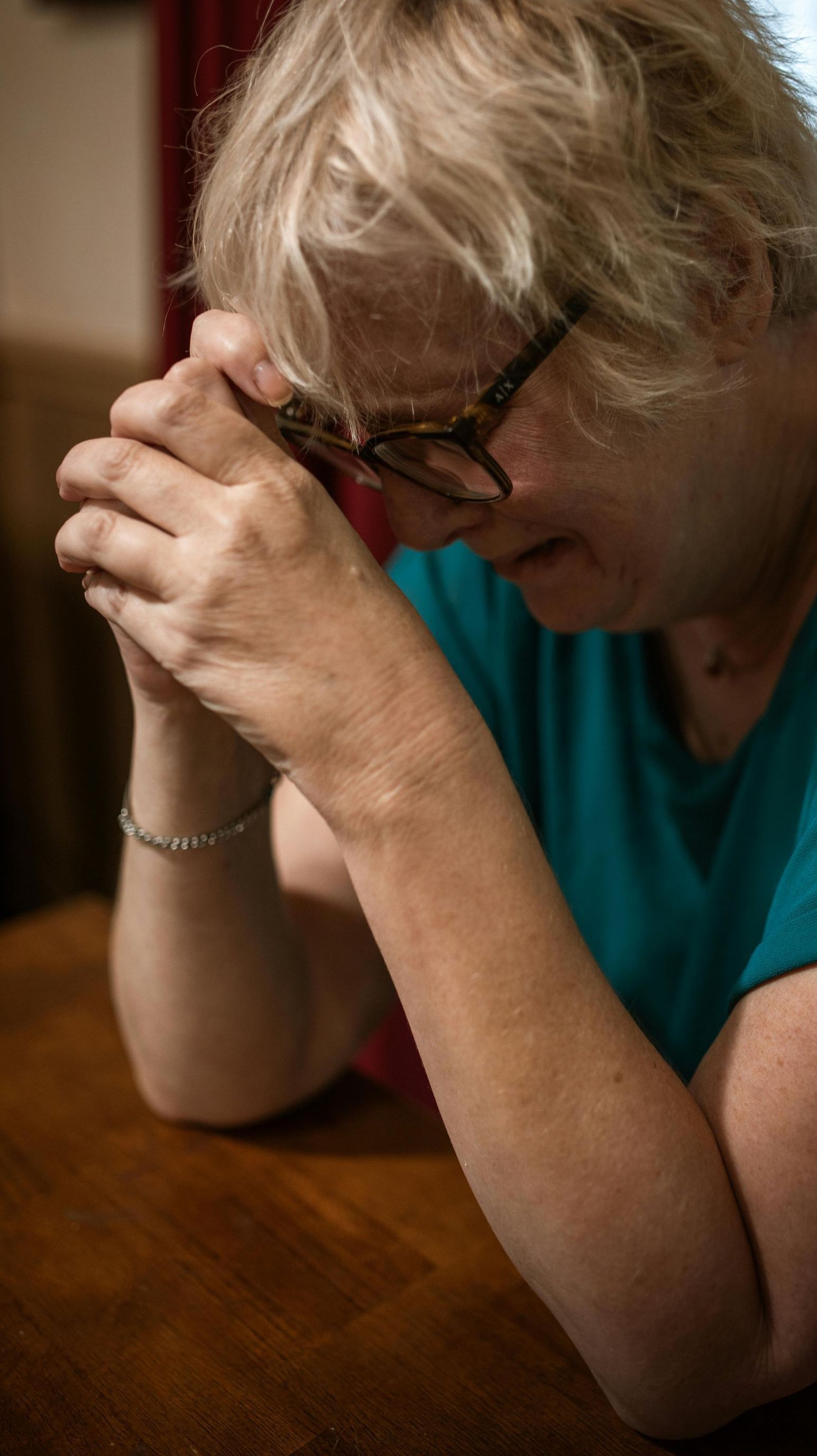 A woman is sitting at a table with her hands folded in prayer.