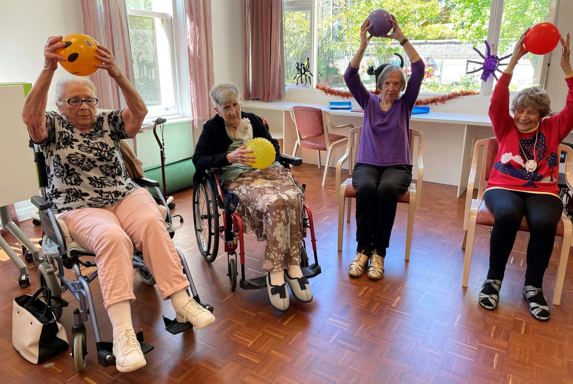 A group of elderly women in wheelchairs are doing exercises with balls.