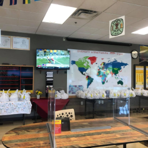 A room with tables holding numerous white gift bags, a world map on the wall, and a television playing a sports game.
