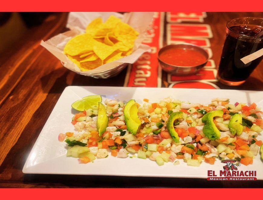 A plate of ceviche topped with avocado slices, served with chips, salsa, and a dark drink on a wooden table.